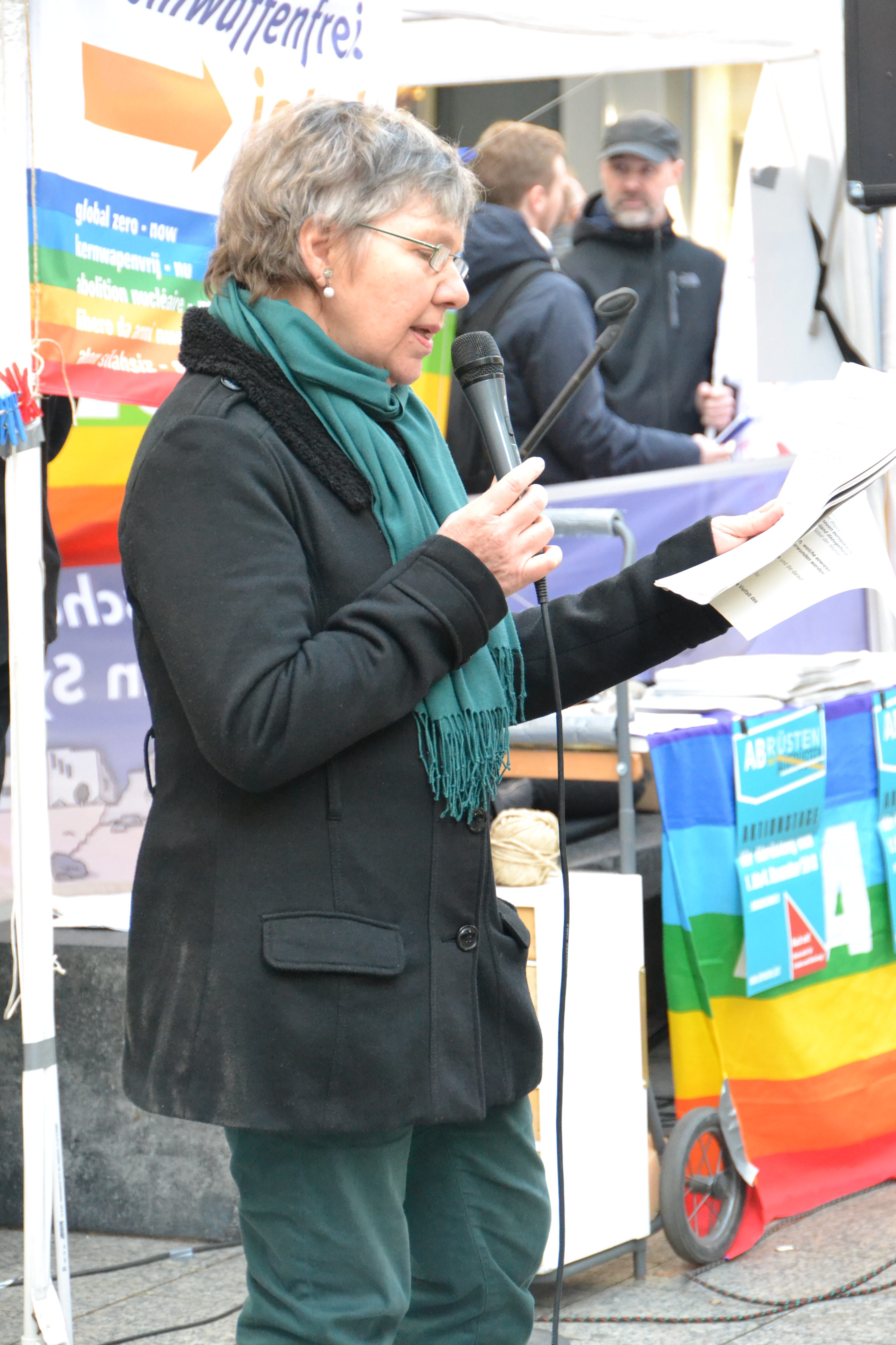 Im Hintergrund sind bunte Friedensfahnen, Kampagnenplakate und Menschen zu sehen.