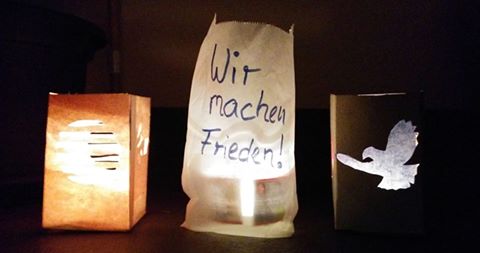 Drei transparente Papiertüten, die als Teelichthalter dienen und Friedenssymbole und -aufschriften zeigen, leuchten vor nachdunklem Hintergrund.