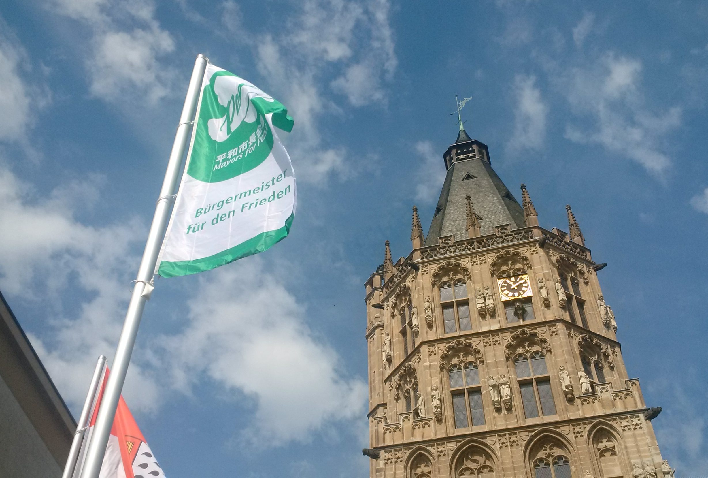 Die Mayors-for-Peace-Flagge trägt eine grüne Aufschrift auf weißem Grund. Hier flattert sich vor dem Kölner Rathausturm. Das Foto ist von unten aufgenommen, sodass man den blauen Himmel und weiße Wölkchen im Hintergrund sieht.