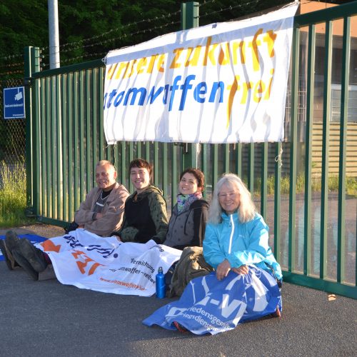 Vier Aktivisten sitzen vor einem Tor, an dem ein Banner mit der Aufschrift "Atomwaffenfrei" hängt. Sie haben sich weitere Banner über die Knie gelegt lachen in die Kamera. Von hinten scheint die aufgehende Sonne durchs Tor.