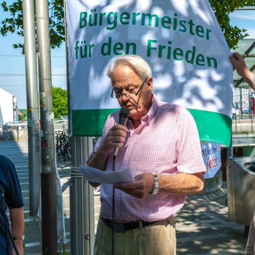 Im Vordergrund stehen zwei Mitglieder der Friedensinitiative Köln-Mülheim in dunkelblauen Vereins-T-Shirts, der Bezirksbürgermeister Norbert Fuchs und Harald Fuchs vom Hiroshima-Nagasaki-Arbeitskreis im Kölner Friedensforum. Im Hintergrund ist der untere Teil der Friedensfahne zu sehen: "Bürgermeister für den Frieden" in grüner Schrift auf weißem Grund.