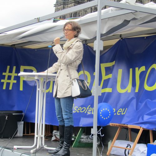 Im Vordergrund steht die Rednerin mit dem Mikro in der Hand auf einem Pritschenwagen. Dahinter sieht man das blaue Transparent mit der gelben Schrift "#PulseOfEurope". Ganz oben am Rand erkennt man ein bisschen vom Turm des Kölner Doms.