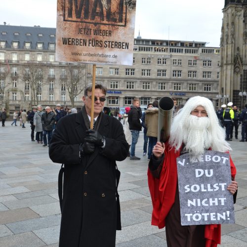 Ein Mann im dunklen Mantel trägt ein Schild im Wüsten-Design der Bundeswehrwerbung mit der Aufschrift "Jetzt wieder werben fürs Sterben!". Neben ihm geht ein bewaffneter Moses mit weißem Rauschebart und einer umgehängten "Steintafel" mit dem 5. Gebot "Du sollst nicht töten!"