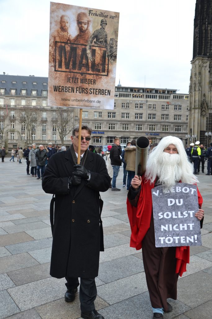 Ein Mann im dunklen Mantel trägt ein Schild im Wüsten-Design der Bundeswehrwerbung mit der Aufschrift "Jetzt wieder werben fürs Sterben!". Neben ihm geht ein bewaffneter Moses mit weißem Rauschebart und einer umgehängten "Steintafel" mit dem 5. Gebot "Du sollst nicht töten!"