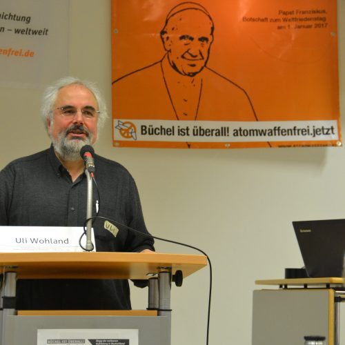 Uli Wohland ist ein Mann mit weißen Haaren und weißgrauem Vollbart, der in einem dunklen Hemd hinter einem Rednerpult steht und spricht. Im Hintergrund ist ein Werbeplakat der Kampagne Atomwaffenfrei jetzt! zu sehen, auf dem Papst Franziskus abgebildet ist.
