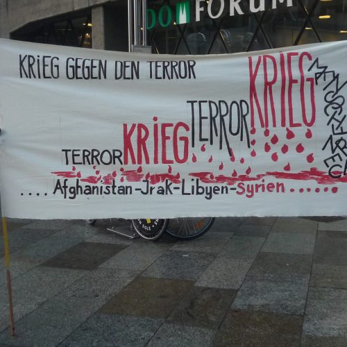 Das selbst auf hellen Stoff gemalte Transparent stellt den "Krieg gegen den Terror" als sich steigernde Abfolge von Krieg und Terror dar. Die Eskalation der Gewalt wird durch sich steigernde Schriftgröße der beiden Wörter, die in rot und schwarz gezeichnet sind, symbolisiert. Darunter regnen Blutstropfen herab, und unten sind die Ländernamen aufgeführt: Afghanistan, Irak, Libyen, Syrien, ...
