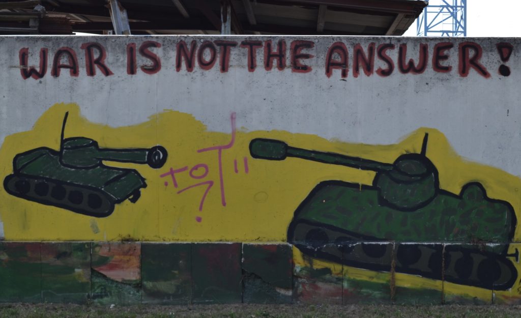 Zwei auf eine Wand gemalte olivgrüne Panzer richten ihre Kanonen aufeinander. Darüber steht in roter Schrift: "War is not the answer!"