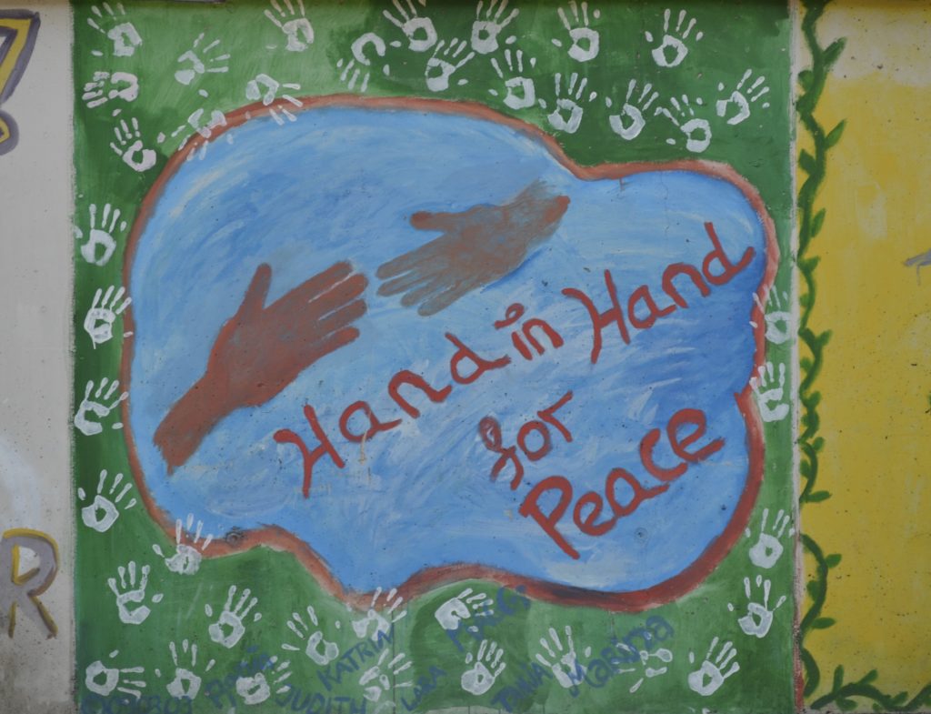 Auf grünem Hintergrund, der mit weißen Handabdrücken bedeckt ist, sieht man in einer Art blauer Wolke zwei rötlich gefärbte Hände, die sich fast berühren, als wollten sich zwei Menschen gerade die Hand geben. Darunter steht "Hand in Hand for Peace".