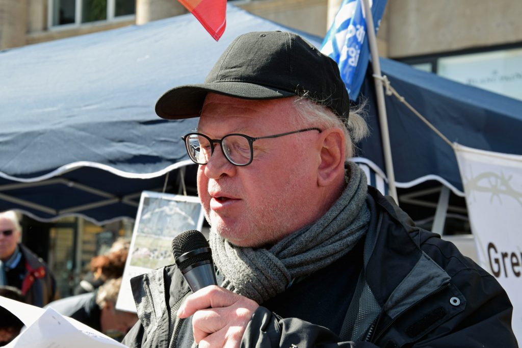 Ein älterer, etwas korpulenter Mann mit dunkler Baseballkappe und Brille spricht ins Mikro. Die Sonne scheint, im Hintergrund sind Transparente und Flaggen, darunter die blauweiße DFG-VK-Flagge, zu erkennen.