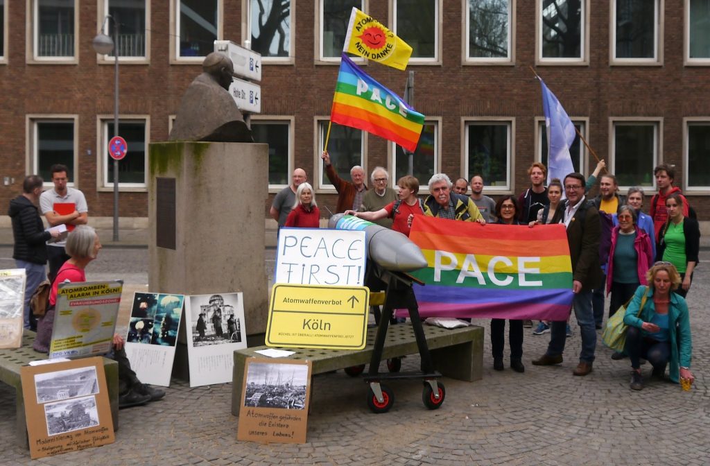 Rund um das steinerne Denkmal von Kardinal Frings sind eine Reihe von Friedensaktivisten mit Plakaten und Friedensfahnen zu sehen. Dazwischen ragt eine graue Atombombe - ein Modell der B61-12 - aus der Gruppe.