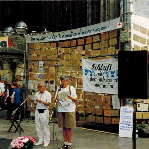 Ein kleiner asiatisch aussehender Mann in weißer Sommerhose und weißem Hemd und eine Frau in T-Shirt und Bermuda-Shorts lesen Texte und sprechen in ein Mikrophon. Sie stehen am Hauptportal des Kölner Doms. Im Hintergrund eine Wand aus lauter Pappschildern, die beschriftet wurden. Darüber hängt ein Transparent mit der Aufschrift "Your signature is a step to elimination of nuclear weapons!" Ein weiteres Transparent mit der Aufschrift "Schluss mit dem nuklearen Wahnsinn" hängt an der Wand. Links sind Zuhörer zu sehen.
