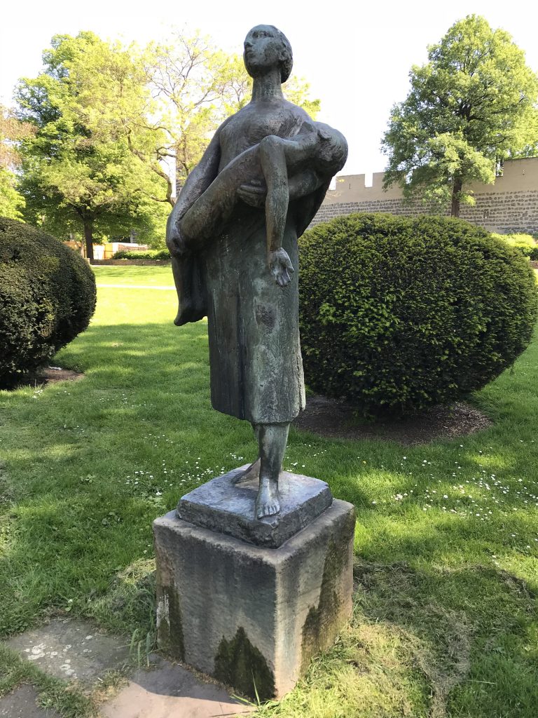 Bronzeskulptur einer Frau, die einen toten Jungen im Arm trägt. Im Hintergrund sind Parkbäume und ein Teil der Kölner Stadtmauer zusehen.
