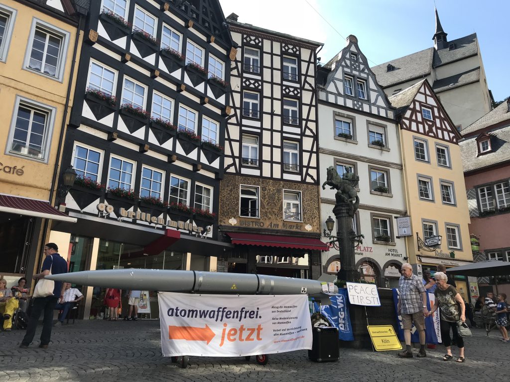 Im Hintergrund sind die prächtigen Fachwerkfassaden von Bürgerhäusern, die einen Markplatz umfassen. Der Brunnen in der Mitte wird teilweise durch eine davor auf einem Gestell liegende hellgraue torpedoförmige Atombombenattrappe verdeckt. An ihr hängt ein Transparent mit der Aufschrift "Atomwaffenfrei jetzt!"