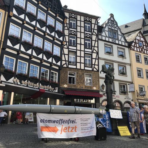 Im Hintergrund sind die prächtigen Fachwerkfassaden von Bürgerhäusern, die einen Markplatz umfassen. Der Brunnen in der Mitte wird teilweise durch eine davor auf einem Gestell liegende hellgraue torpedoförmige Atombombenattrappe verdeckt. An ihr hängt ein Transparent mit der Aufschrift "Atomwaffenfrei jetzt!"