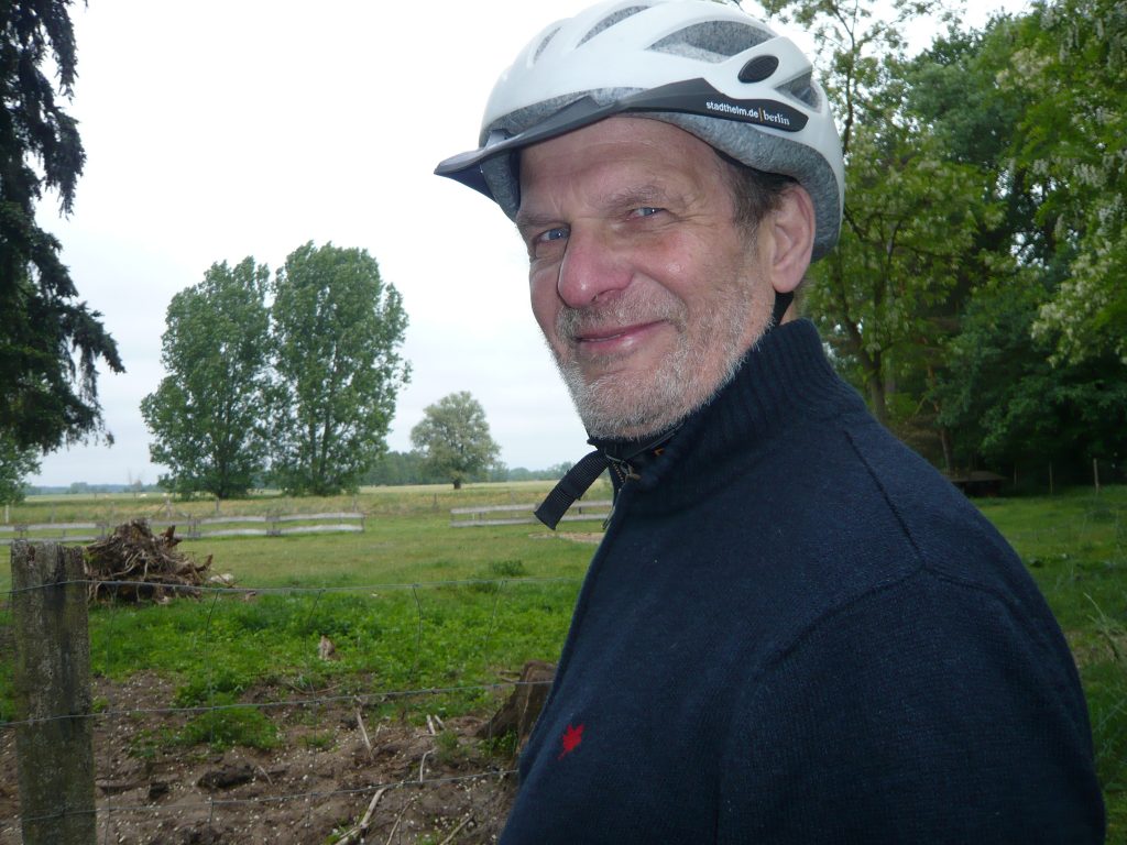 Ein älterer Herr mit kurzem hellgrauen Bart und blauen Augen, mit einem hellen Fahrradhelm auf dem Kopf und in dunklem Pulli schaut freundlich in die Kamera. Im Hintergrund ist eine Landschaft mit Feldern und Bäumen.