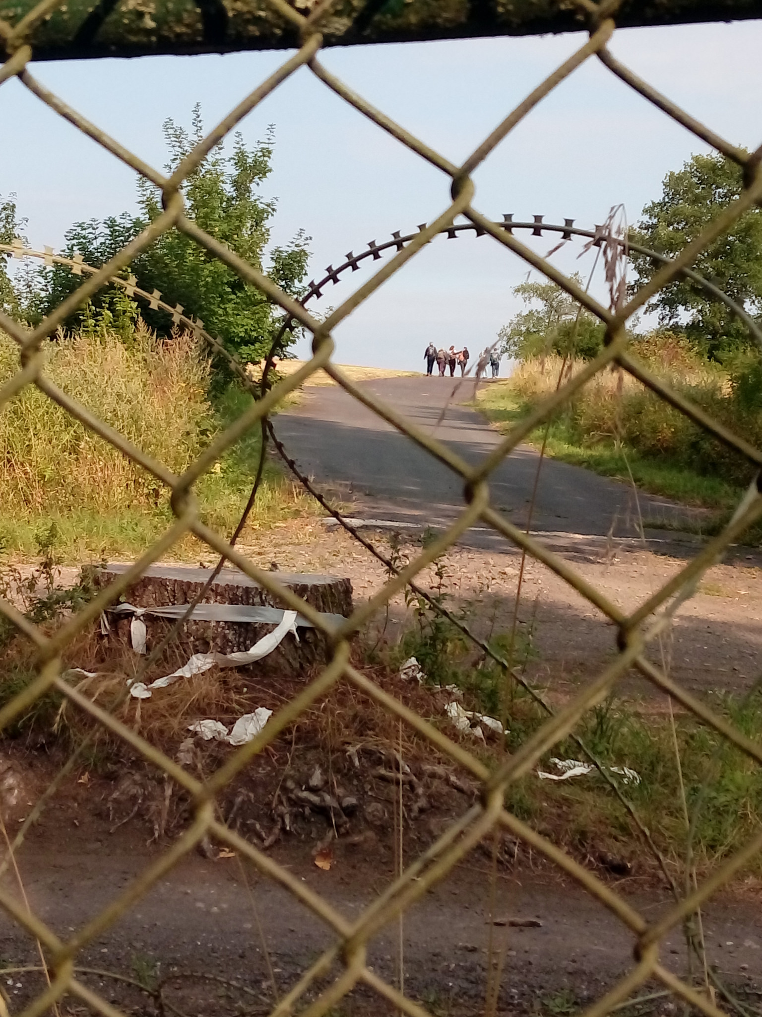 Durch einen Maschendraht und einen NATO-Stacheldraht hindurch sieht man einen Weg, der zwischen Büschen und trockenem Gras einen Hügel hinaufführt. Oben am Ende des Wegs zeichnen sich ein paar Menschen gegen den blauen Himmel ab.