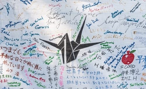 Ausschnitt eines mit vielen deutschen, japanischen und anderen Schriftzügen beschriebenes weißes Transparent. In der Mitte der gemalte Origami-Kranich.