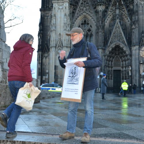Ein älterer schlanker Herr mit einem Sandwich-Plakat vor der Brust wendet sich einer Passantin zu und übergibt ihr einen Flyer. Im Hintergrund ein Teil des Hauptportals des Kölner Doms.