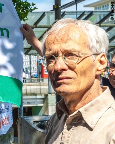 Ein älterer Mann schaut skeptisch am Fotografen vorbei. Er hat ein sommerlich gebräuntes Gesicht und schüttere hellgraue Haare.