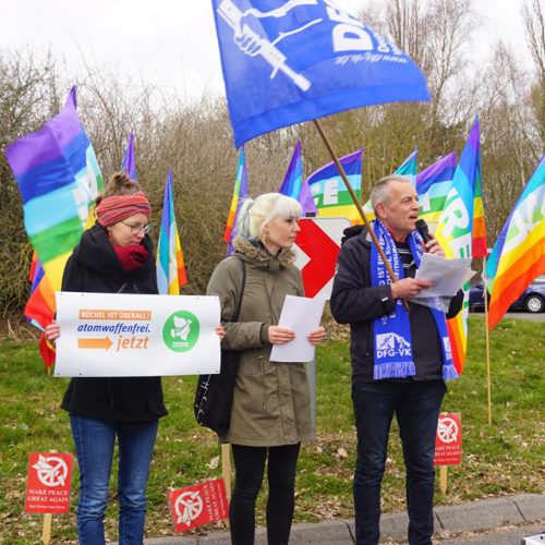 Zwei Frauen und ein Mann stehen winterlich gekleidet nebeneinander; im Hintergrund zahlreiche bunt gestreifte Friedensfahnen und die blauweiße Fahne der DFG-VK.
