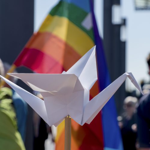 Im Vordergrund ein weißer Origami-Papierkranich auf einem Stock und dahinter bunte Friedensfahnen im Sonnenlicht.