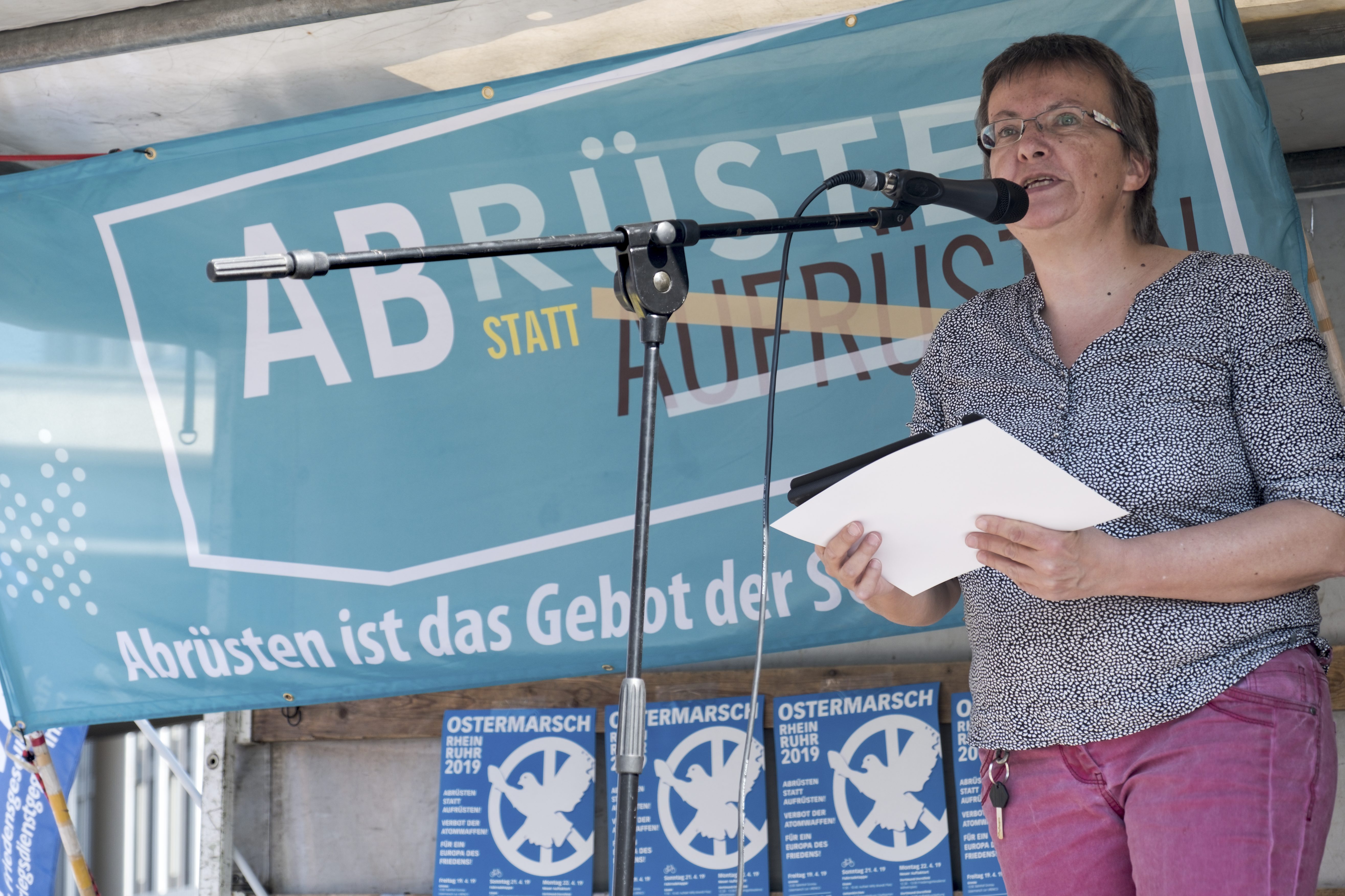 Eine sportlich aussehende ältere Dame mit sehr kurzgeschnittenen braunen Haaren und Brille, in grauem T-Shirt und rosa Jeans, spricht auf einer improvisierten Bühne auf einem LKW in ein Mikro. Im Hintergrund ein türkisfarbenes Transparent "Abrüsten statt Aufrüsten" und blau-weiße Ostermarsch-Plakate.