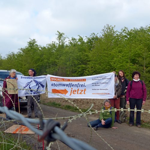 Eine Gruppe hält auf einer Fahrpiste Transparente "Frieden schaffen ohne Waffen" und "Büchel ist überall - Atomwaffenfrei jetzt!". Im Vordergrund sind konventioneller und NATO-Stacheldraht zu sehen.
