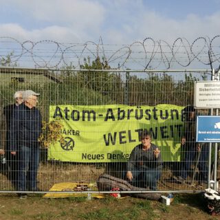 Hinter einem stacheldrahtbewehrten Bauzaun sitzt eine ältere Dame neben einer Gitarre und einer kleinen Picknickdecke auf dem Boden. Drei ältere Herren stehen neben der Decke und tragen einen dicken Strauß gelb blühender Ginsterzweige. Hinter der Dame hängt an einem weiteren Drahtzaun ein großes gelbes Transparent "Atom-Abrüstung weltweit. Neues Denken. Quäker"