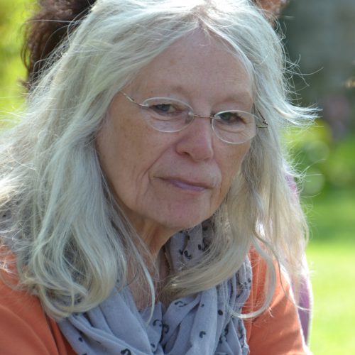 Eine nachdenklich-freundliche ältere Dame mit schönen Gesichtszügen und dicken, schulterlangen, in der Mitte gescheitelten weißgrauen Haaren, blaugrauem Halstuch und orangefarbenem Shirt vor einem sonnig-frühlingshaften Wiesenhintergrund.