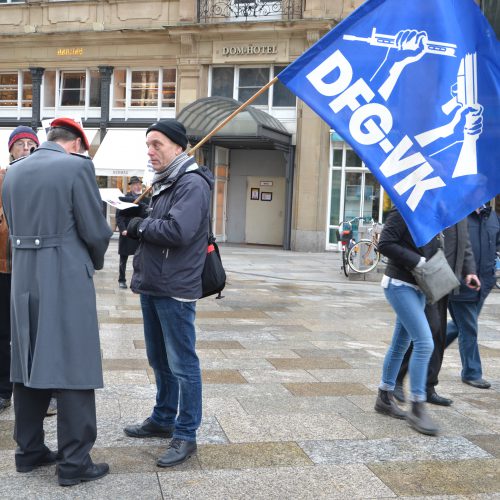 Ein älterer Mann mit dunkler Mütze und einer blau-weißen DFG-VK-Fahne mit dem zerbrochenen Gewehr in der Hand spricht mit einem Soldaten in einem langen grauen Mantel und einer weißroten Mütze.