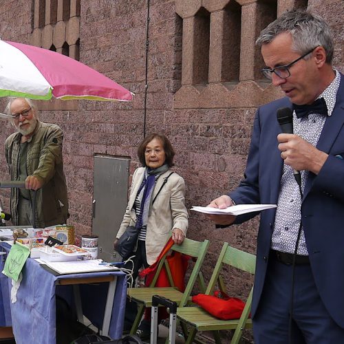 Ein schlanker Mann mit Brille und graumelierten Haaren spricht in ein Mikro. Er trägt einen blauen Anzug und Fliege. Links von ihm hören eine kleine asiatisch aussehende Dame und ein älterer Herr mit weißem Bart zu. Sie stehen an einem kleinen Tisch mit Infomaterial und grünen Holzstühlen.