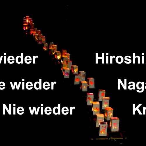 Auf fast schwarzem, leicht reflektierendem Wasser treibt eine lockere Doppelreihe von weißen Papierlaternchen mit ausgeschnittenen Muster, durch die rötlich die brennenden Kerzen schimmern. Links und rechts daneben steht mit weißer Schrift "Nie wieder Hiroshima. Nie wieder Nagasaki. Nie wieder Krieg."