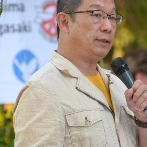 Ein asiatisch aussehender Herr mit kurzen schwarzen Haaren und Brille, in einem beigefarbenen Leinenhemd und sonnengelbem T-Shirt darunter spricht in ein Mikrophon. Im Hintergrund ein Schild mit einer Friedenstaube und dem Logo von ICAN, der Internationalen Kampagne zur Abschaffung der Kernwaffen.