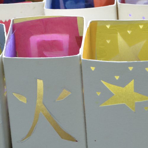 Eine Reihe weißer Papierlaternchen mit ausgeschnittenen Mustern (Kölner Dom, ein japanisches Schriftzeichen, Sternchen), die mit buntem Transparenpapier beklebt sind.