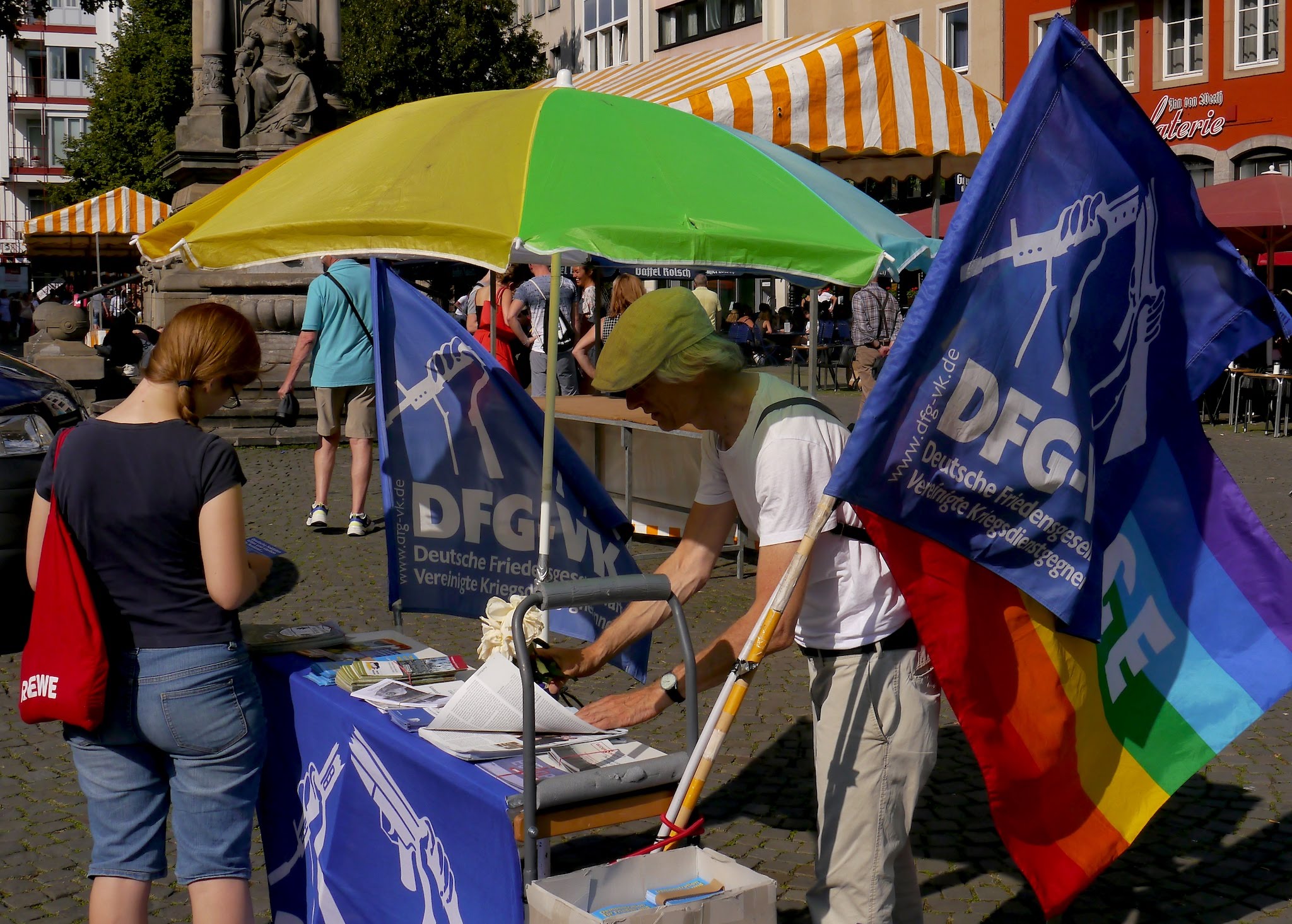 Ein schlanker älterer Herr mit Kappe in weißem T-Shirt und heller Sommerhose schreibt etwas auf einem mit Dokumenten bedeckten und blauen DFG-VK-Fahnen ausstaffierten Tischchen. Ein bunter Sonnenschirm sorgt für Schatten.