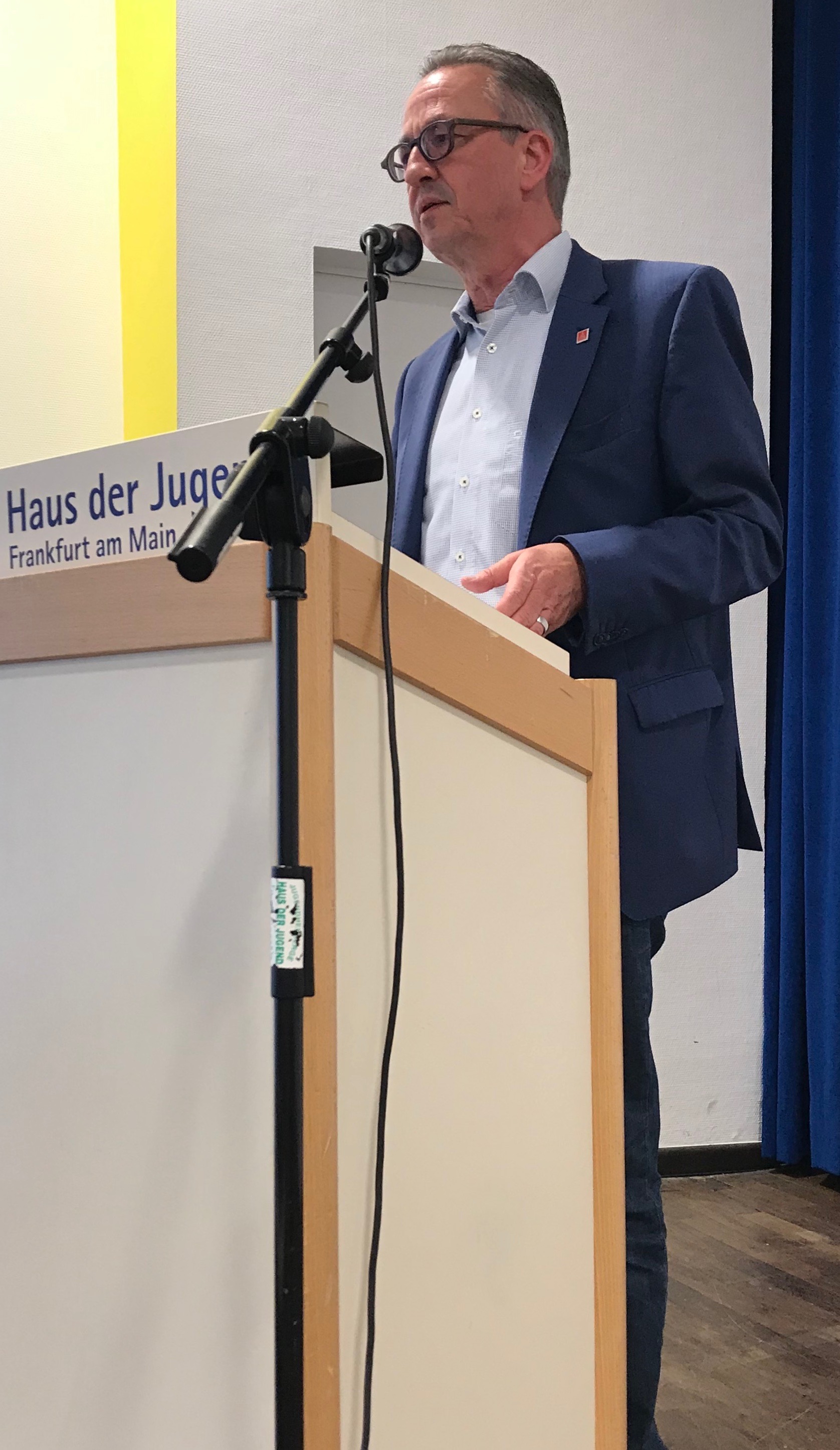 Ein mittelalter Herr in dunklem Anzug, weißem Hemd steht hinter einem hellen Rednerpult und spricht in ein Mikro.