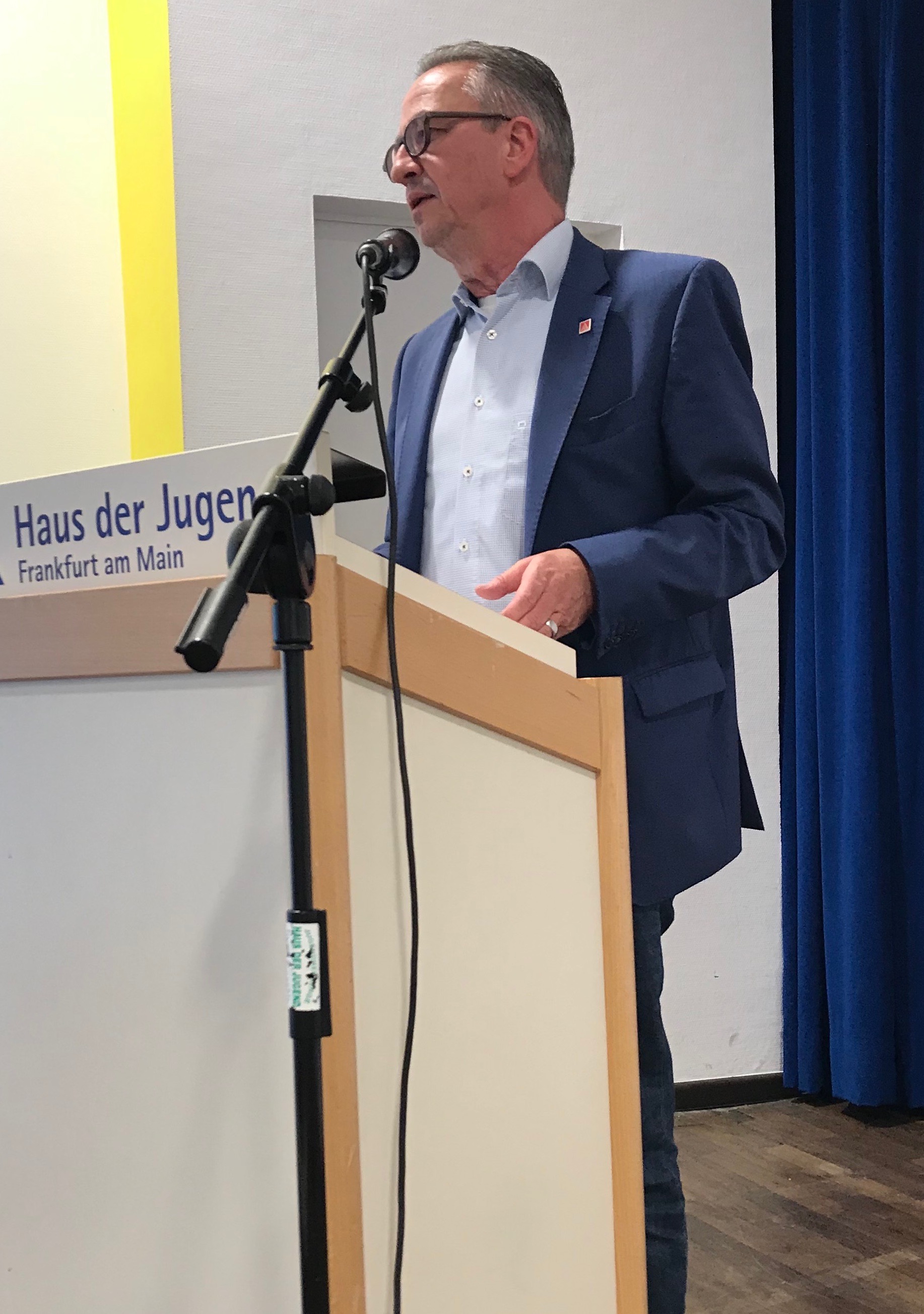 Ein mittelalter Herr in dunklem Anzug, weißem Hemd steht hinter einem hellen Rednerpult und spricht in ein Mikro.