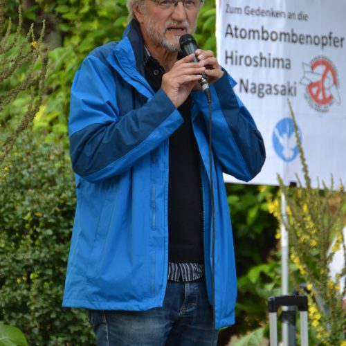 Ein schlanker älterer Herr in blauer sportlicher Jacke und Jeans spricht in ein Mikro. Im Hintergrund ein Transparent mit der Aufschrift "Zum Gedenken an die Atombombenopfer", "Hiroshima", "Nagasaki"