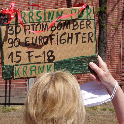 Eine Frau mit blondem Kurzhaarschnitt, die von hinten zu sehen ist, hält ihr selbst beschriftetes Pappschild in die Höhe.