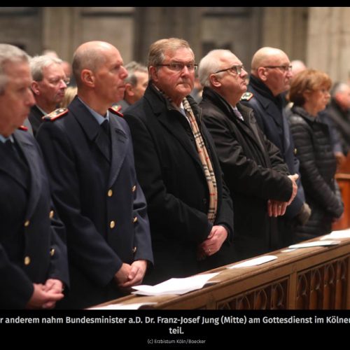 Die erste Reihe im Kirchenschiff zeigt Herren in dunklen Uniformmänteln und Oberbürgermeisterin Reker, die zum Altar schauen. Nur ein Mann im dunklen Zivilanzug schaut nach rechts, direkt ins Objektiv der Kamera.