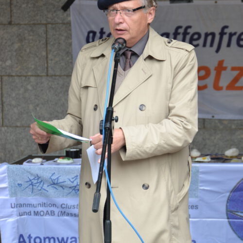 Ein mittelalter Mann mit hellem Trenchcoat und schwarzer Mütze spricht in ein Mikro. Im Hintergrund ist ein Banner mit "atomwaffenfrei Jetzt!" zu erkennen.