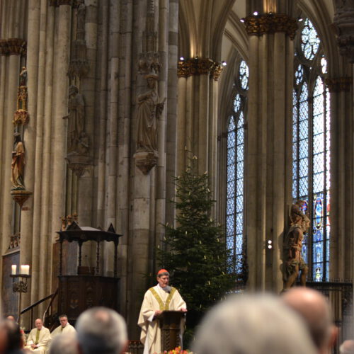 Ein feierlich hell gekleideter Priester mit roter Kopfbedeckung redet an einem Pult. Im Vordergrund die ihm zugewandten Köpfe und Schultern von Männern in Uniformen, die in Bänken sitzen. Im Hintergrund die riesigen Säulen und Fenster eines gotischen Doms und eine weihnachtlich geschmückte Fichte.