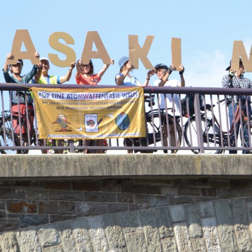 Über ein Dutzend Leute stehen hinter einem Brückengeländer und halten Pappbuchstaben hoch. Am Geländer hängen Transparente mit den Aufschriften "Atomwaffenfrei jetzt!" und "Für eine atomwaffenfreie Welt!"
