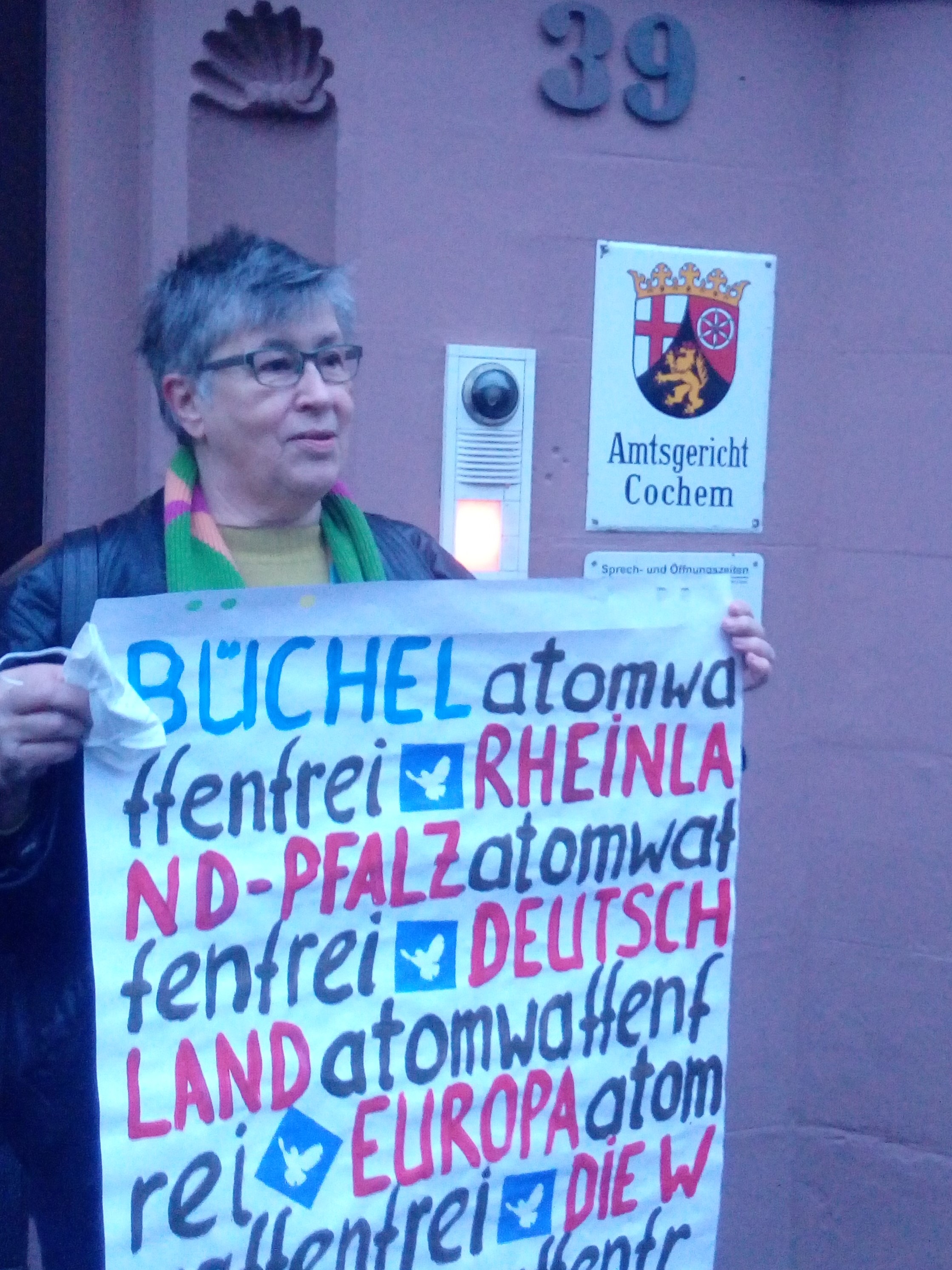 Eine ältere Frau mit kurzen grauen Haaren hält, vor dem Amtsgerichtsgebäude Chochem stehend, ein selbst gemaltes DIN A2-Papier vor sich.