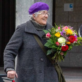 Eine alte Dame in dunkelgrauem Wollmantel und lilafarbener Filzmütze stützt sich leicht auf den Griff eines Rollators, während sie strahlend einen fröhlich-bunten Blumenstrauß in der Hand hält.