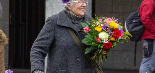 Eine alte Dame in dunkelgrauem Wollmantel und lilafarbener Filzmütze stützt sich leicht auf den Griff eines Rollators, während sie strahlend einen fröhlich-bunten Blumenstrauß in der Hand hält.