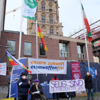 Eine ältere Dame sitzt im Rollator und spricht in das Mikro eines Megafons, das von einem Helfer gehalten wird. Rechts im Bild Transparente "Selig sind die Frieden stiften", "unsere zukunft atomwaffenfrei", "nuclear weapons are banned", sowie die Flaggen Deutschlands, Nordrhein-Westfalens und der "Mayors for peace", vor dem Turm des Historischen Rathauses der Stadt Köln.