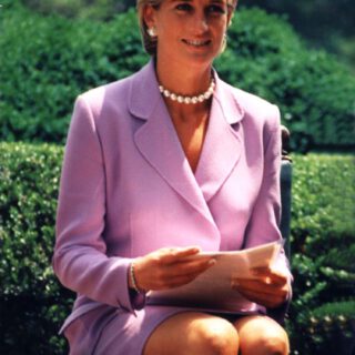 Eine junge Frau mit kurzen blonden Haaren, Perlenohrsteckern und einem Perlencollier in einem rosafarbenem Kostüm hält Papier in den Händen, während sie auf einem Hocker in einem sonnigen Park sitzt.