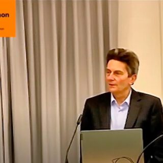 Ein Mann mit dichten schwarzen Haaren in dunklem Jackett und hellem Hemd spricht stehend hinter einem aufgeklappten Laptop in ein Mikro.