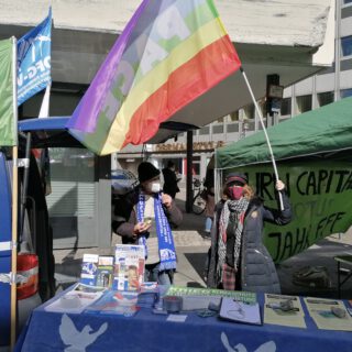 Ein Mann und eine Frau stehen in Winterkleidung und Stoffmasken hinter einem Stand, der mit Friedenstauben auf blauem Grund bedeckt ist und auf dem Flyer liegen. Die Frau schwenkt eine große Friedensfahne. Im Hintergrund weitere Fahnen und Pavillons.